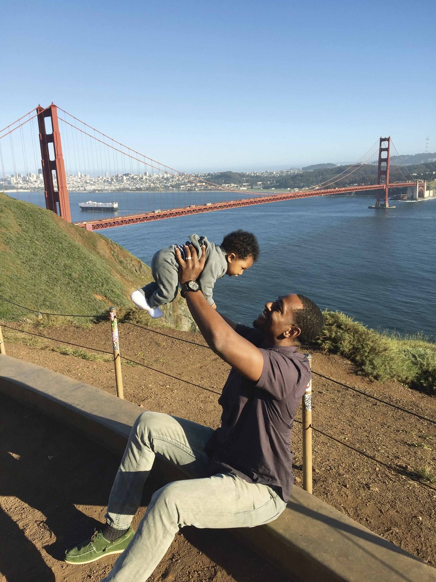 dad holds infant up in the air, smiling at the baby, in front of the Golden Gate Bridge