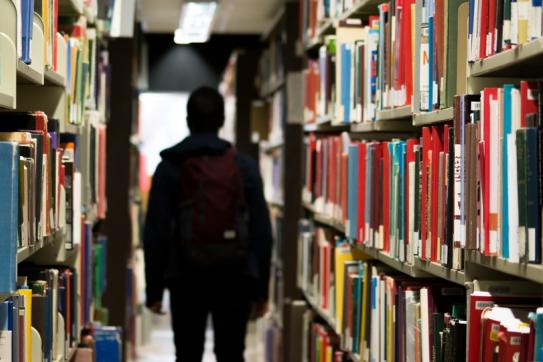 Student in a backpack walking between 2 shelves of library books