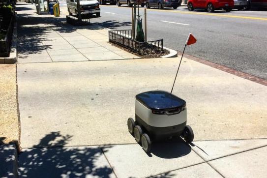 A mobile delivery robot trundles along the sidewalk